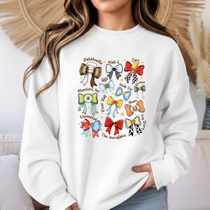 May include: White sweatshirt featuring colorful bow designs inspired by various Pixar movies, including "Ratatouille," "Cars," and "Toy Story." Each bow is uniquely designed with movie-related elements and text. The sweatshirt has a crew neck and long sleeves.