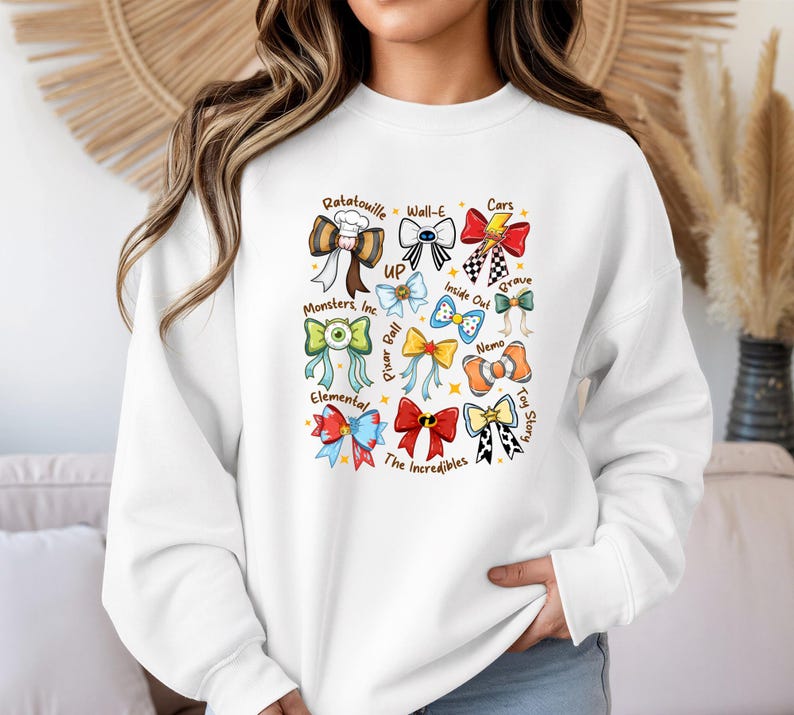 May include: White sweatshirt featuring colorful bow designs inspired by various Pixar movies, including "Ratatouille," "Cars," and "Toy Story." Each bow is uniquely designed with movie-related elements and text. The sweatshirt has a crew neck and long sleeves.