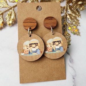 May include: A pair of wooden earrings with a cartoon image of an elderly couple holding mugs. The earrings are brown with a wood grain pattern and have silver hooks.