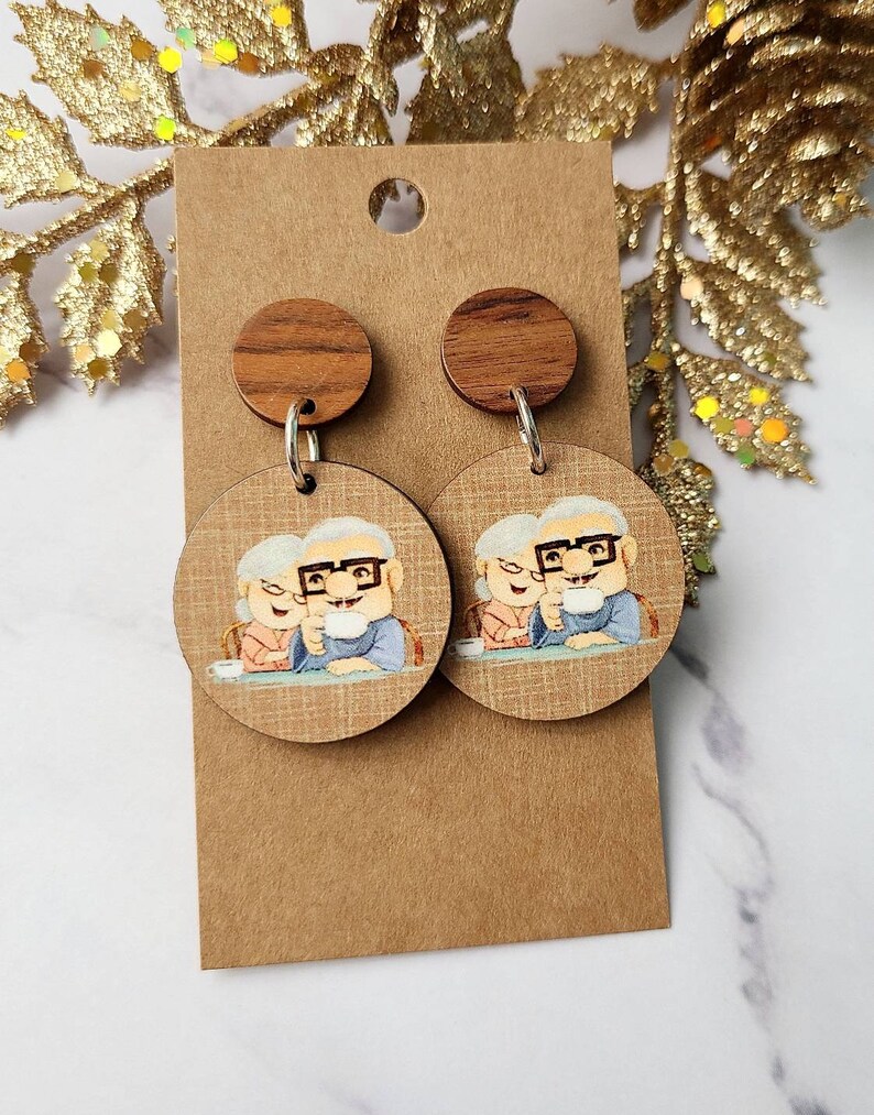 May include: A pair of wooden earrings with a cartoon image of an elderly couple holding mugs. The earrings are brown with a wood grain pattern and have silver hooks.