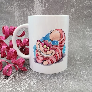 May include: White ceramic mug with a colorful illustration of the Cheshire Cat from Alice in Wonderland. The cat is grinning with a wide, toothy smile and has pink and red stripes.