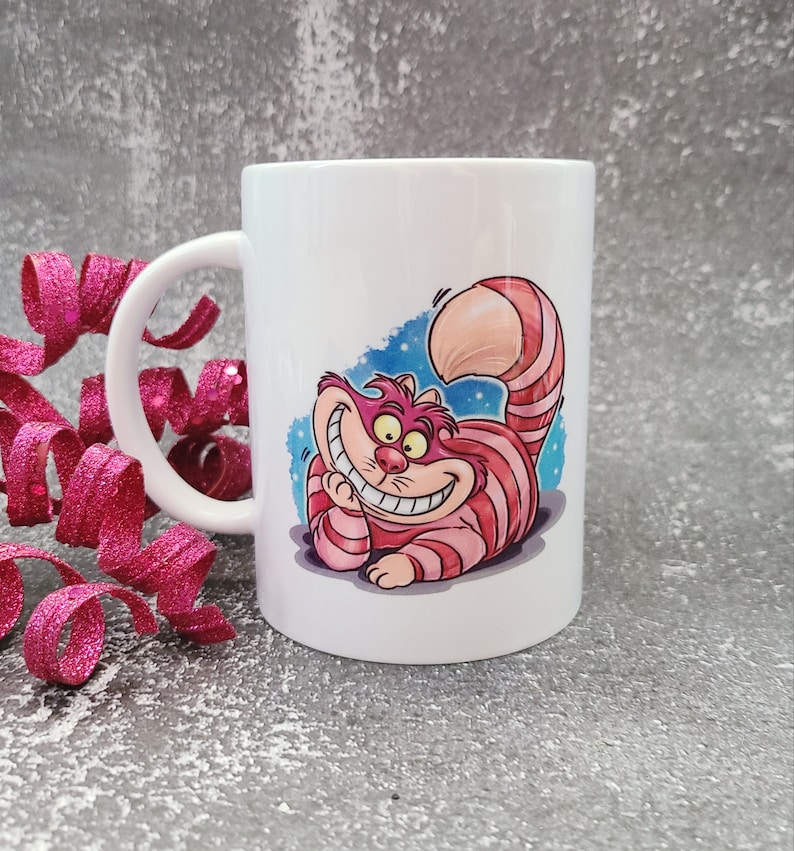 May include: White ceramic mug with a colorful illustration of the Cheshire Cat from Alice in Wonderland. The cat is grinning with a wide, toothy smile and has pink and red stripes.