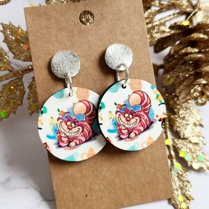 May include: A pair of round dangle earrings featuring the Cheshire Cat from Alice in Wonderland. The earrings have a silver stud and a circular pendant with a colorful illustration of the cat. The background is white with colorful accents.