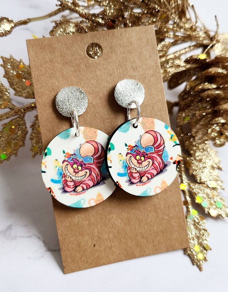 May include: A pair of round dangle earrings featuring the Cheshire Cat from Alice in Wonderland. The earrings have a silver stud and a circular pendant with a colorful illustration of the cat. The background is white with colorful accents.