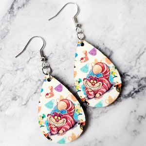 May include: A pair of teardrop-shaped earrings featuring the Cheshire Cat from Alice in Wonderland. The earrings have a white background with colorful teacups and the Cheshire Cat's smiling face. The earrings are made of a lightweight material and have silver-colored ear wires.