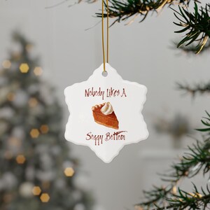 Pode incluir: Enfeite de Natal branco em forma de floco de neve com o texto "Nobody Likes A Soggy Bottom" em castanho. O enfeite apresenta uma ilustração em aquarela de uma fatia de torta com chantilly. Uma corda dourada é anexada para pendurar.