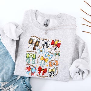 May include: A light gray sweatshirt featuring colorful bow designs inspired by various animated movies, including "Ratatouille," "Cars," and "Toy Story." The bows are arranged in a grid pattern, with each bow representing a different film. The sweatshirt has a crew neck and long sleeves.
