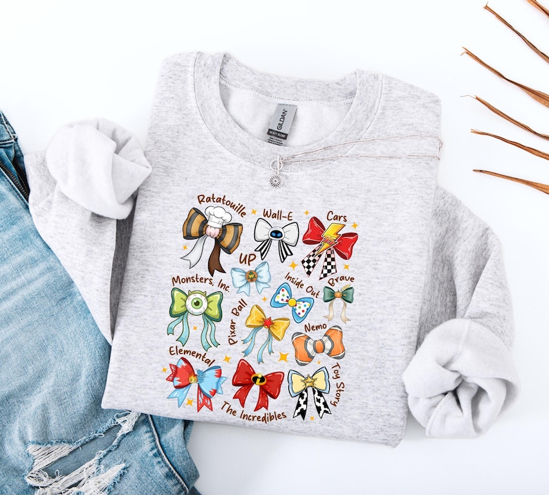 May include: A light gray sweatshirt featuring colorful bow designs inspired by various animated movies, including "Ratatouille," "Cars," and "Toy Story." The bows are arranged in a grid pattern, with each bow representing a different film. The sweatshirt has a crew neck and long sleeves.