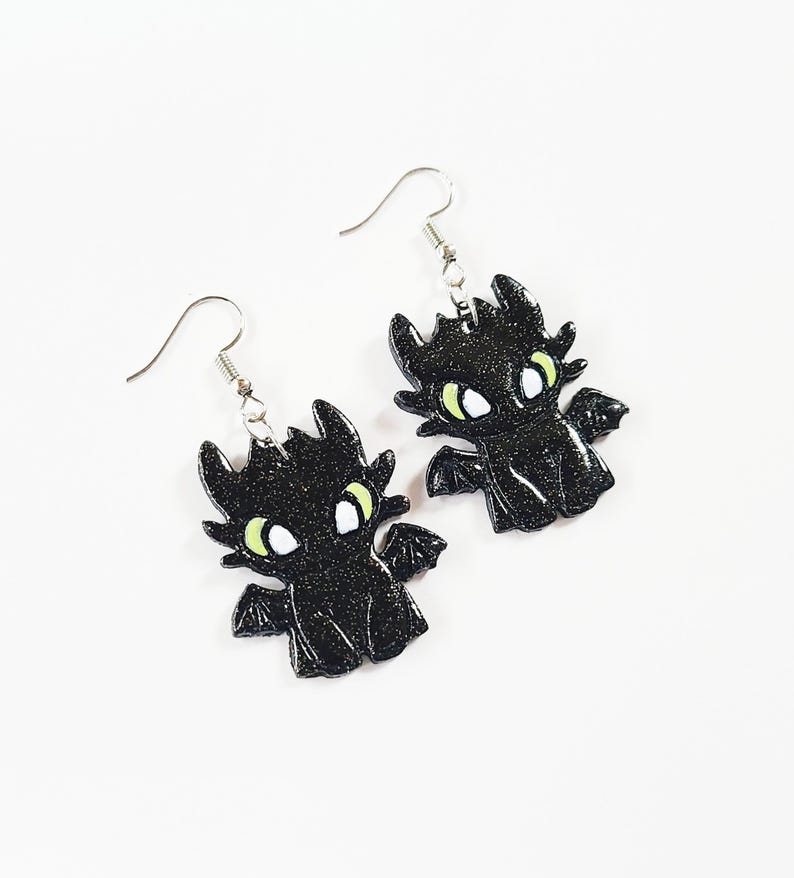 May include: A pair of black glitter dragon earrings with green eyes and white pupils. The earrings are on a white background.