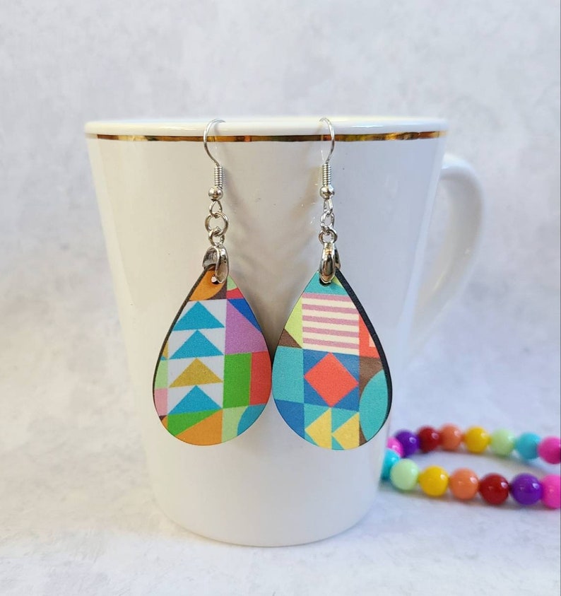 May include: A pair of teardrop-shaped earrings with a colourful geometric pattern. The earrings are made of wood and have silver hooks.