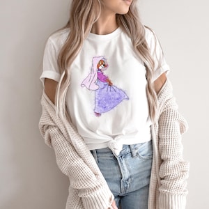 Op de afbeelding: Wit T-shirt met een cartoonillustratie van een vos die een paarse jurk en een roze sluier draagt. De vos kijkt naar rechts en heeft een roze blos op zijn wangen.