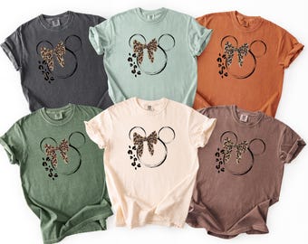 May include: Six t-shirts in different colours, each with a black outline of Mickey Mouse's head, a leopard print bow, and paw prints. The shirts are in shades of grey, green, light blue, orange, cream, and brown.