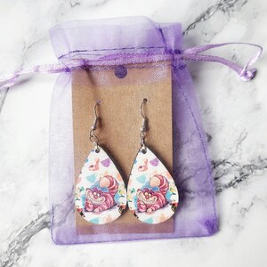 May include: A pair of teardrop-shaped earrings featuring the Cheshire Cat from Alice in Wonderland. The earrings have a white background with a colorful pattern and a smiling pink cat. They are displayed in a purple drawstring bag.