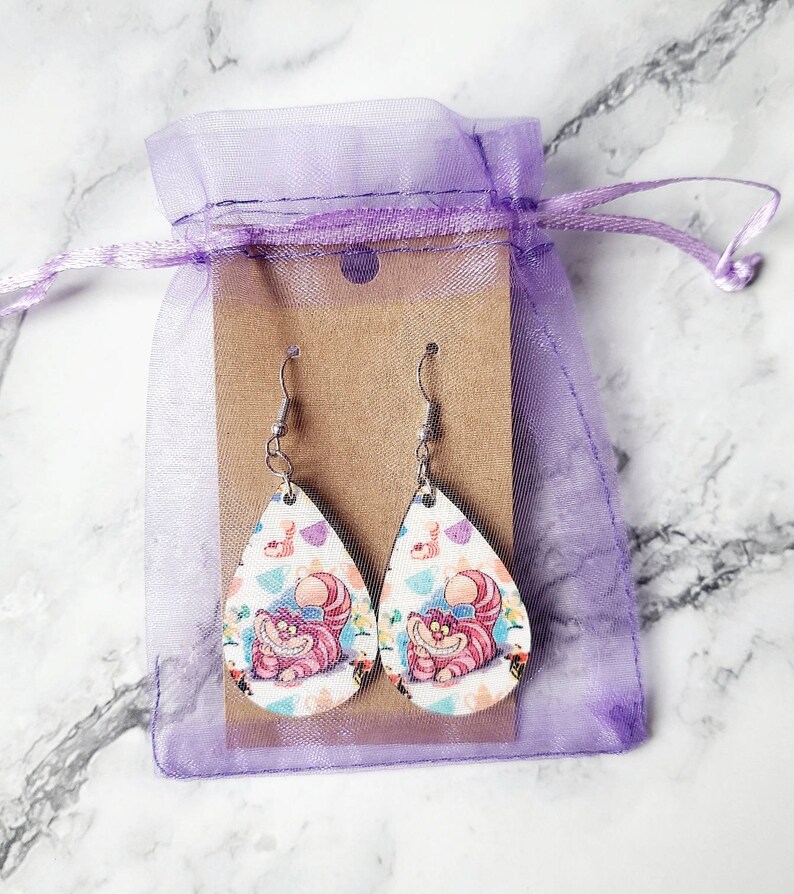 May include: A pair of teardrop-shaped earrings featuring the Cheshire Cat from Alice in Wonderland. The earrings have a white background with a colorful pattern and a smiling pink cat. They are displayed in a purple drawstring bag.