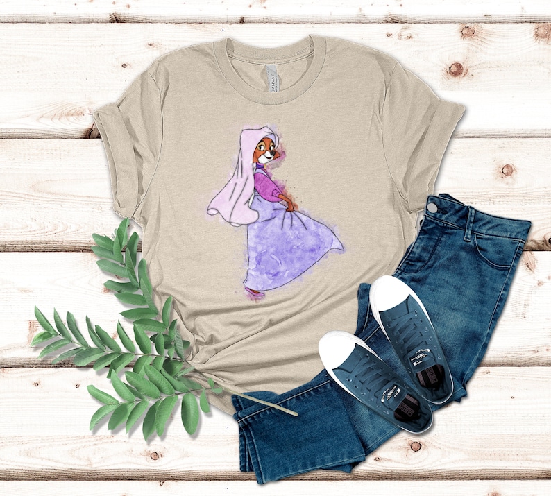 Op de afbeelding: Een beige T-shirt met een aquarel-illustratie van een cartoonvos die een paarse jurk en een witte sluier draagt. De vos kijkt naar rechts en heeft een roze neus en een roze strik op zijn kop.
