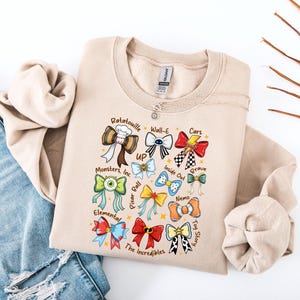May include: Beige sweatshirt featuring colorful bow designs inspired by animated movies like "Ratatouille," "Cars," and "Toy Story." The bows are adorned with movie-related graphics and text, including "Monsters, Inc." and "The Incredibles."