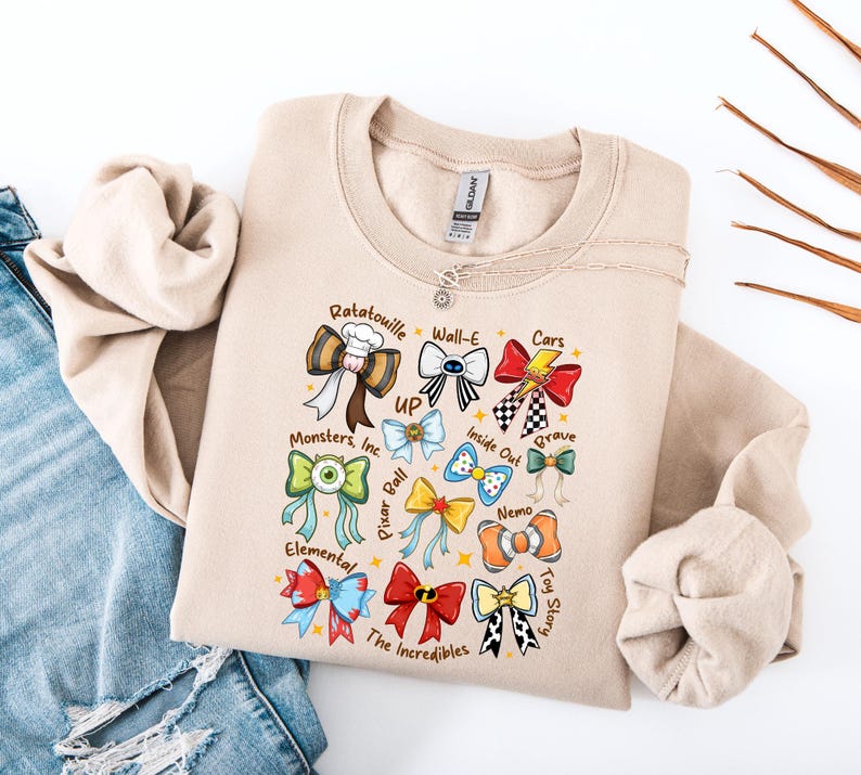 May include: Beige sweatshirt featuring colorful bow designs inspired by animated movies like "Ratatouille," "Cars," and "Toy Story." The bows are adorned with movie-related graphics and text, including "Monsters, Inc." and "The Incredibles."