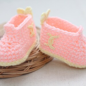 Peut inclure: Une paire de chaussons pour bébé faits à la main. Ces chaussures au crochet sont rose clair avec une bordure et des accents jaune clair. Les chaussons ont un motif décoratif sur le côté et un petit nœud à l'arrière.