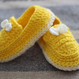 Peut inclure: Une paire de chaussons de bébé au crochet jaunes faits à la main, avec une bordure blanche et de petits nœuds blancs. Les chaussures sont faites de fil doux et sont conçues pour être confortables pour les pieds d'un bébé.
