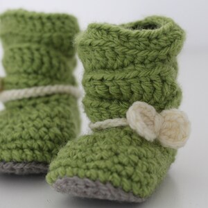 Peut inclure: Une paire de chaussons de bébé au crochet verts faits à la main. Chaque chausson est orné d'un nœud crème et d'un détail de corde crème assorti autour de la cheville. Les semelles sont grises.