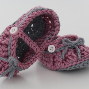 Peut inclure: Une paire de chaussons pour bébé faits à la main au crochet. Les chaussures sont de couleur rose poudré avec des accents gris, notamment les semelles, les bordures et les brides. Chaque chaussure a un petit nœud et une fermeture à bouton.