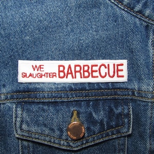 May include: White patch sewn onto a blue denim jacket with red embroidered text that reads "WE SLAUGHTER BARBECUE"
