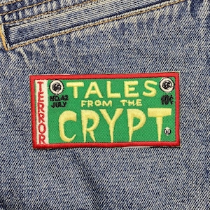 Könnte beinhalten: Ein gestickter Aufnäher in Grün und Rot mit dem Text "Tales from the Crypt" und den Worten "Terror" und "July" links und "10¢" rechts. Der Aufnäher ist auf eine blaue Jeansjacke genäht.