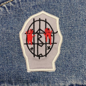 Könnte beinhalten: Eine graue Jeansjacke mit einem roten und schwarzen gestickten Aufnäher. Der Aufnäher zeigt eine Hand mit einem Käfig darum.