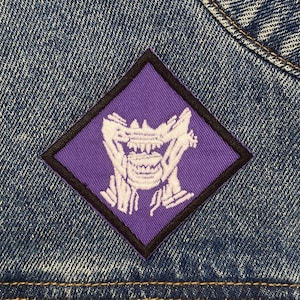 May include: Purple diamond-shaped patch with a white embroidered design of a creature with a wide mouth and teeth. The patch is sewn onto a blue denim jacket.