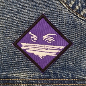Op de afbeelding: Close-up van een blauw spijkerjack met een paarse patch. De patch is ruitvormig met een zwarte rand en heeft een witte afbeelding van ogen en een horizontale lijn over de mond.