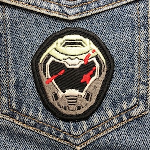 May include: A black and gray embroidered patch of a demon's helmet with red accents on a blue denim jacket.