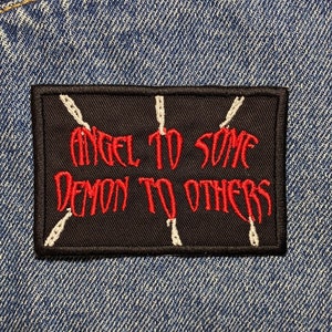May include: A black patch with white stitching and red text that reads "Angel to some Demon to others" on a blue denim background.