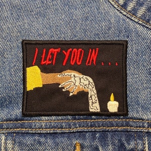 May include: Embroidered patch with the text "I LET YOU IN..." in red on a black background. The patch features a hand with a candle and a yellow sleeve. The patch is sewn onto a denim jacket.