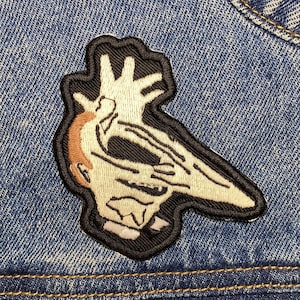 Peut inclure: Un écusson brodé blanc et noir représentant une créature au long nez et une main sur les yeux, cousu sur une veste en jean bleue.