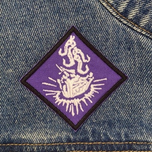 May include: Purple diamond-shaped patch with white embroidered design of a hand holding a heart with flames and a starburst around it.