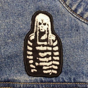 May include: White embroidered patch of a skeleton figure wearing a striped shirt on a blue denim background.