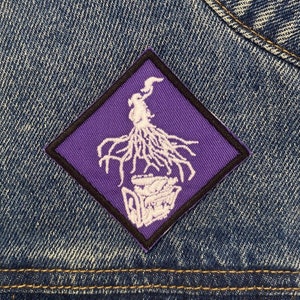 May include: A white embroidered patch with a purple background in the shape of a diamond. The patch features a stylized image of a plant with roots and a pot.