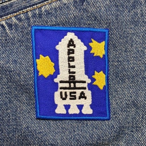 Op de afbeelding: Een blauwe en witte geborduurde patch met een raket en de tekst "Apollo USA". De patch heeft gele sterren rond de raket.