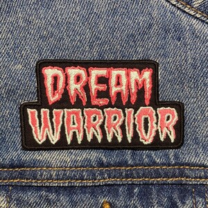 Op de afbeelding: Een close-up van een blauw spijkerjasje met een zwart embleem. Het embleem toont de woorden "DREAM WARRIOR" in een rode, gestileerde letter met een witte omtrek. De jas heeft een koperkleurige knoop.