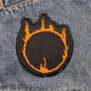 May include: A black and orange embroidered patch with a circle design surrounded by flames. The patch is sewn onto a blue denim jacket.