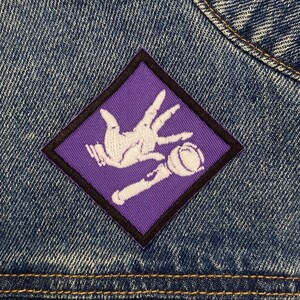 May include: Purple diamond-shaped patch with a white outline. The patch features a white hand holding a white microphone. The hand is making a hand gesture with the index and middle fingers extended and the thumb pointing up.