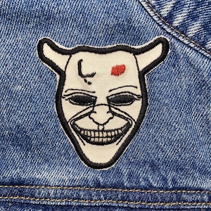 May include: A white embroidered patch with a black outline of a face with horns, a red dot on the forehead, and a wide smile. The patch is sewn onto a blue denim jacket.