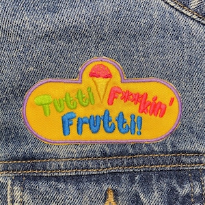 Może przedstawiać: Żółta łatka w kształcie chmury z napisem "Tutti Frutti!" w kolorowych literach. Nad tekstem znajduje się różowy i czerwony rożek lodowy.