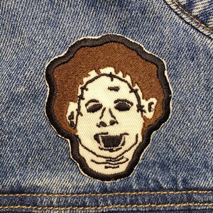 May include: A brown and white embroidered patch of a person's face with a stitched-on mouth and eyes. The patch is sewn onto a blue denim jacket.