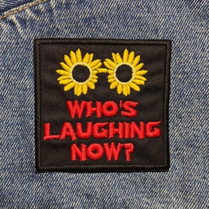 Può includere: Una toppa nera con due girasoli ricamati gialli e il testo "WHO'S LAUGHING NOW?" in ricamo rosso. La toppa è cucita su una giacca di jeans blu.