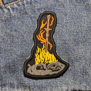 Może przedstawiać: Czarna haftowana łatka z mieczem w ogniu, otoczona czaszkami. Miecz jest srebrny, a ogień jest żółty i pomarańczowy.