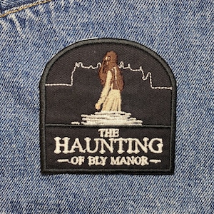 May include: A black and white embroidered patch featuring a silhouette of a woman walking in front of a building. The text "THE HAUNTING - OF BLY MANOR -" is below the image.