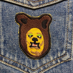 Peut inclure: Un écusson brodé représentant un visage d'ours brun avec une fourrure jaune, des yeux verts et une bouche rose. L'écusson est cousu sur une veste en jean bleue.