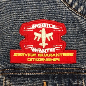 Op de afbeelding: Rode en gele geborduurde patch met de tekst "MOBILE INFANTRY SERVICE GUARANTEES CITIZENSHIP". De patch is bevestigd aan een blauwe denimjas.