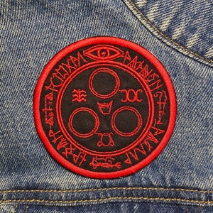 Peut inclure: Patch brodé rouge avec un fond noir représentant un cercle avec trois petits cercles à l'intérieur et un symbole au-dessus. Le cercle est entouré d'un anneau de texte qui dit "AGNOSTI-CISM" en cercle.